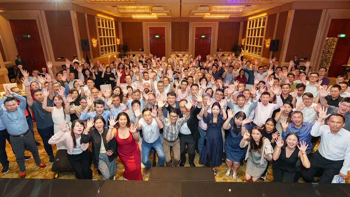 A large company group filling a ballroom at an annual dinner, arms raised for one wide group photograph