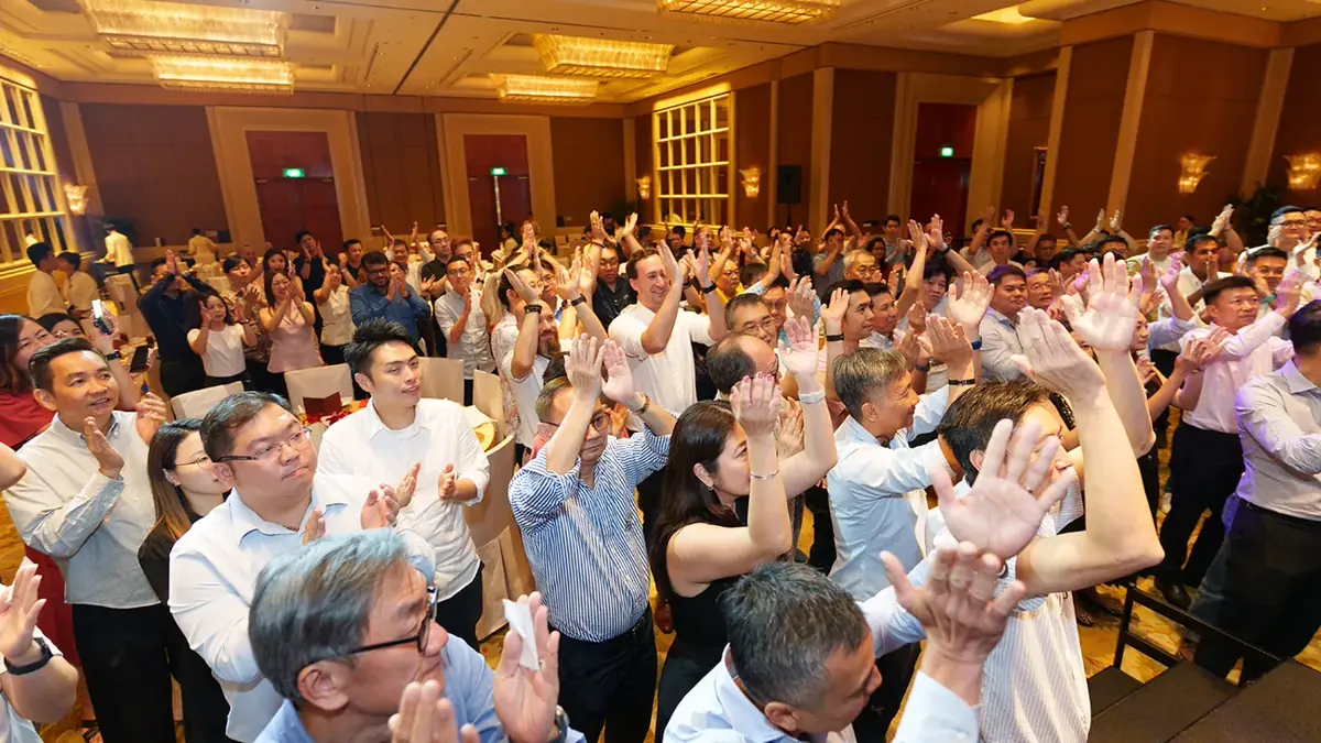 A corporate event venue in Singapore in use: a hotel ballroom packed with guests standing and applauding under warm chandeliers, banquet chairs and a pillar behind them