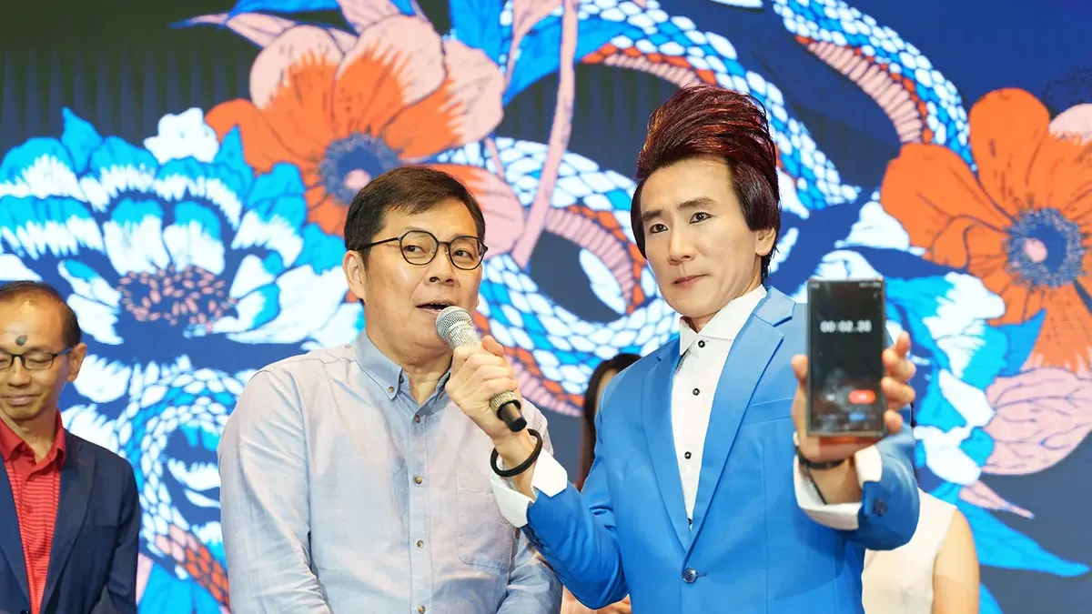Two men at the front of a stage before a large floral screen, one speaking into a microphone and one in a blue suit holding up a phone with a timer running, with three more people partly in view beside and behind them
