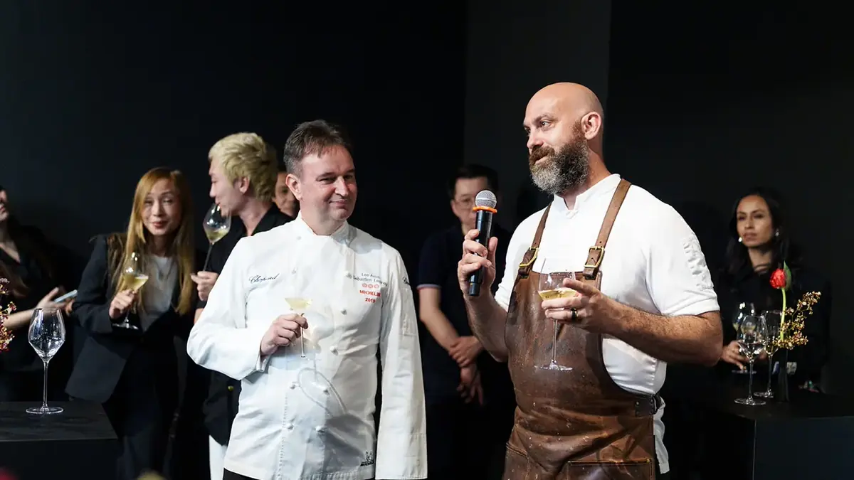 Two chefs addressing guests holding wine glasses at a small private company dinner, in warm low light