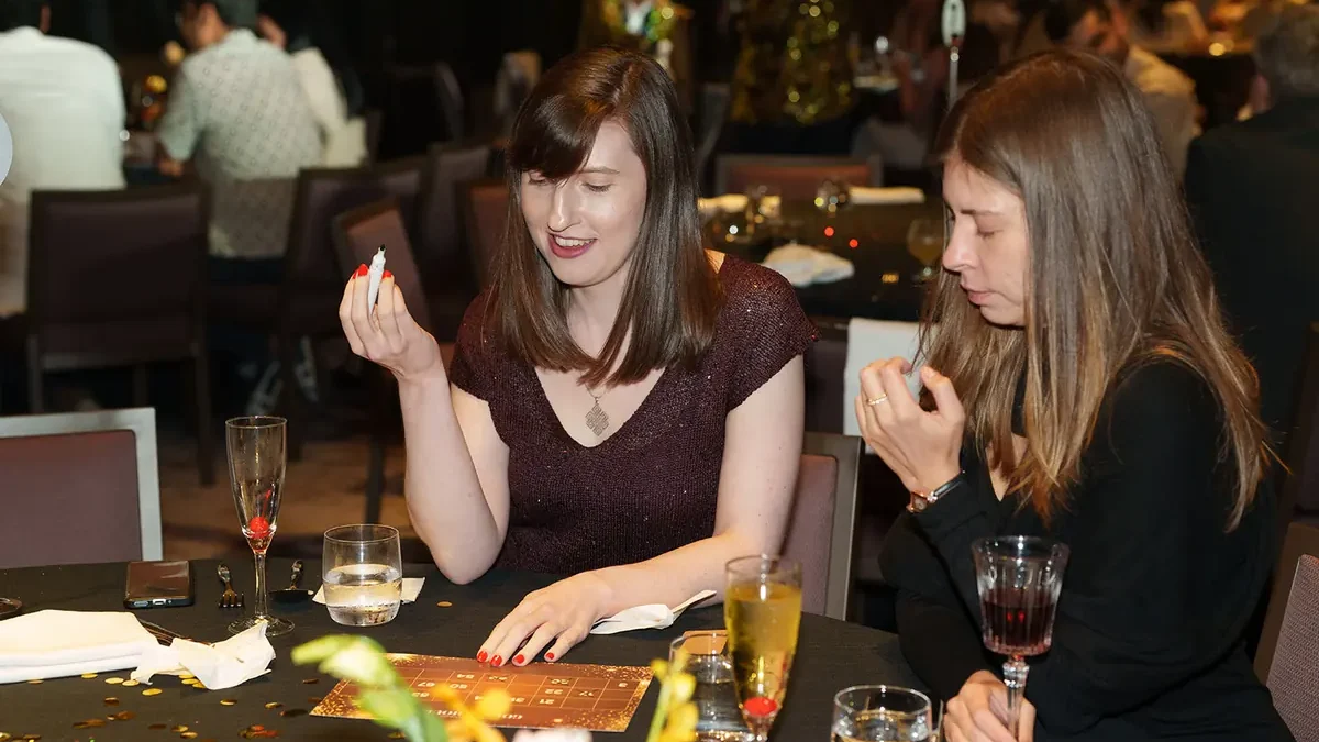 Two guests at a dinner table, one holding up a marker with her other hand on a printed grid card, glasses and gold confetti on the tablecloth