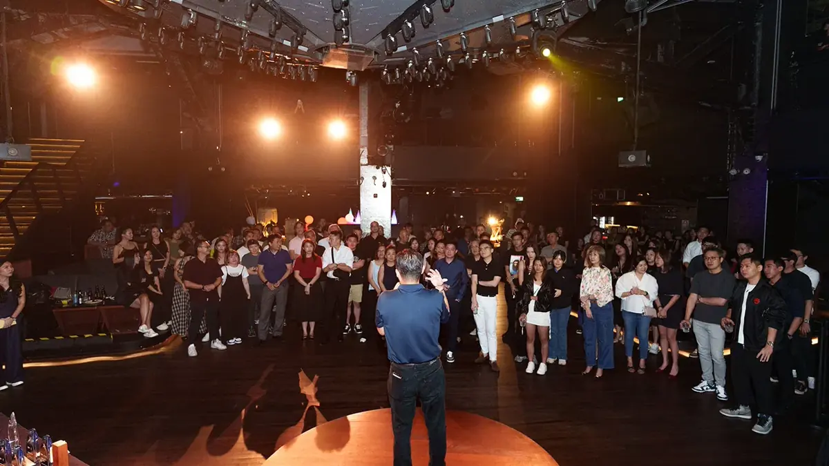 Guests standing in a darkened event venue around a central lit stage with one person on it