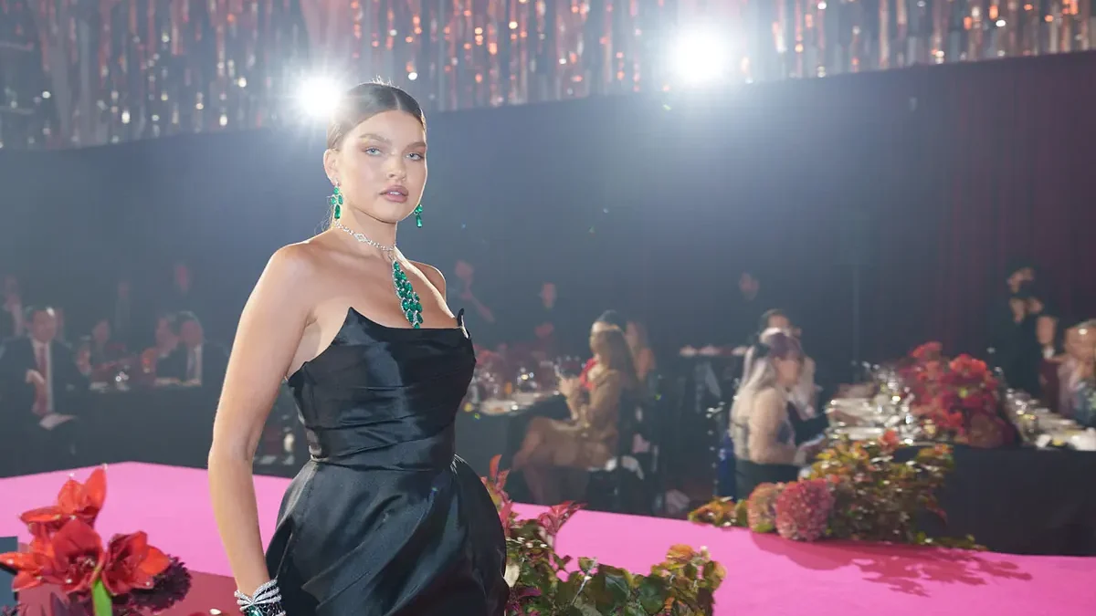 A model in a black gown and green jewellery on a pink runway edged with flowers at a gala dinner, with guests seated at their tables behind her under a ceiling of hanging lights