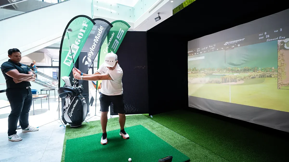 A golfer mid-swing on a strip of turf inside a black-walled simulator bay in an atrium, with a projection screen across the back wall and three tall feather flags at its open front