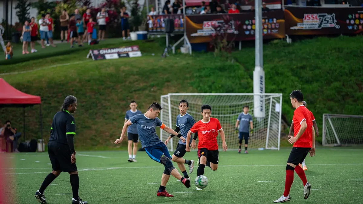 A football match on an artificial pitch, with a red canopy tent at the touchline and spectators on the grass bank above