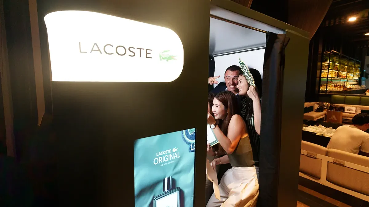 A photo booth with a brand name on a lit sign above a screen, three guests posing inside its open side
