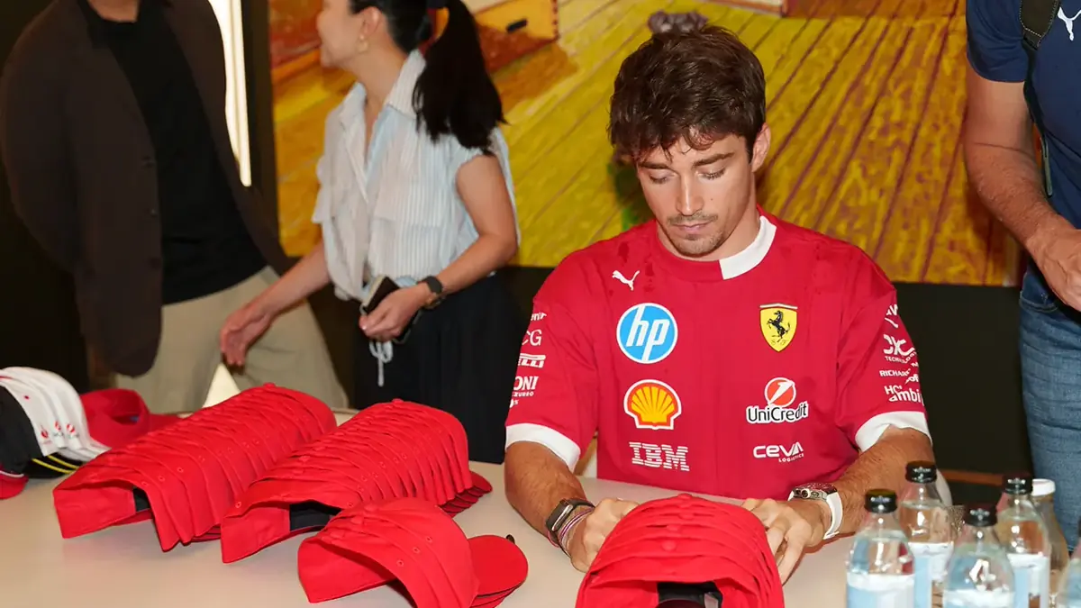 A meet-the-driver signing session on a branded trade show stand, visitors gathered at a table stacked with red caps