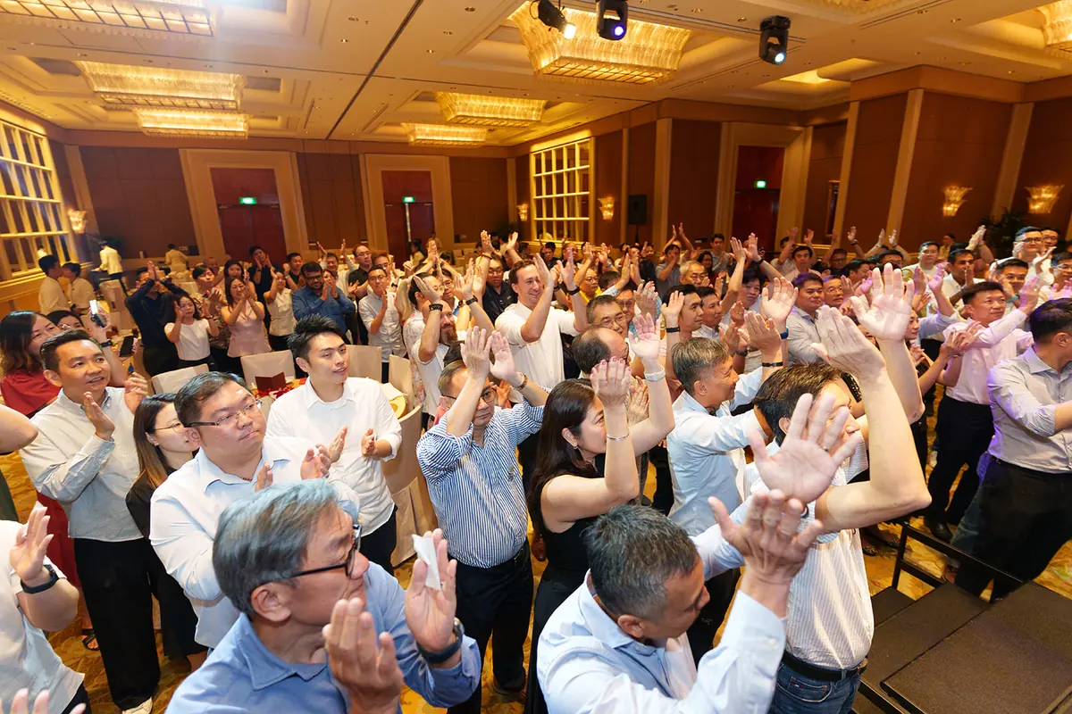 A ballroom of guests in business attire stand clapping and raising their hands beneath chandeliers