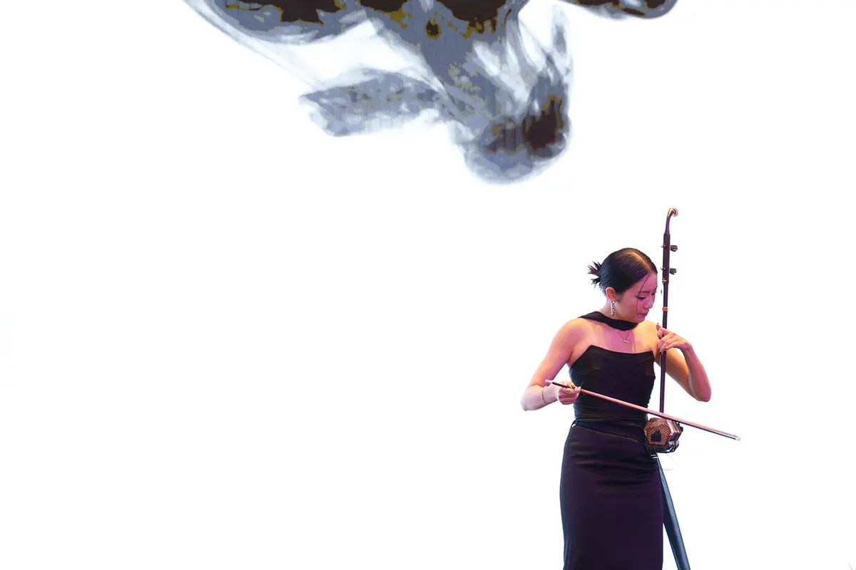 A musician in a black strapless dress plays an erhu on stage against a bright white screen