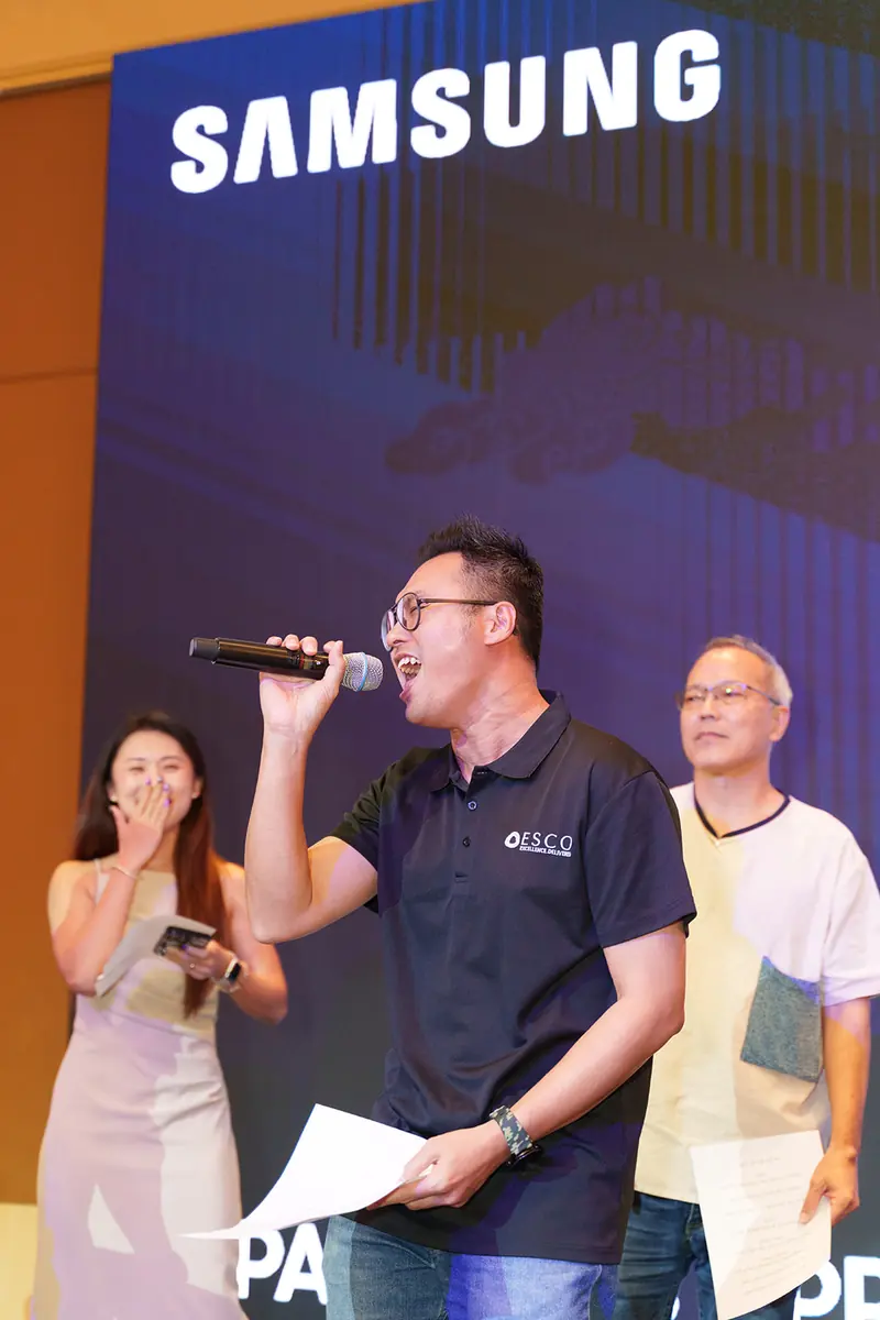 A guest in a dark polo sings into a microphone on stage while a woman beside him laughs