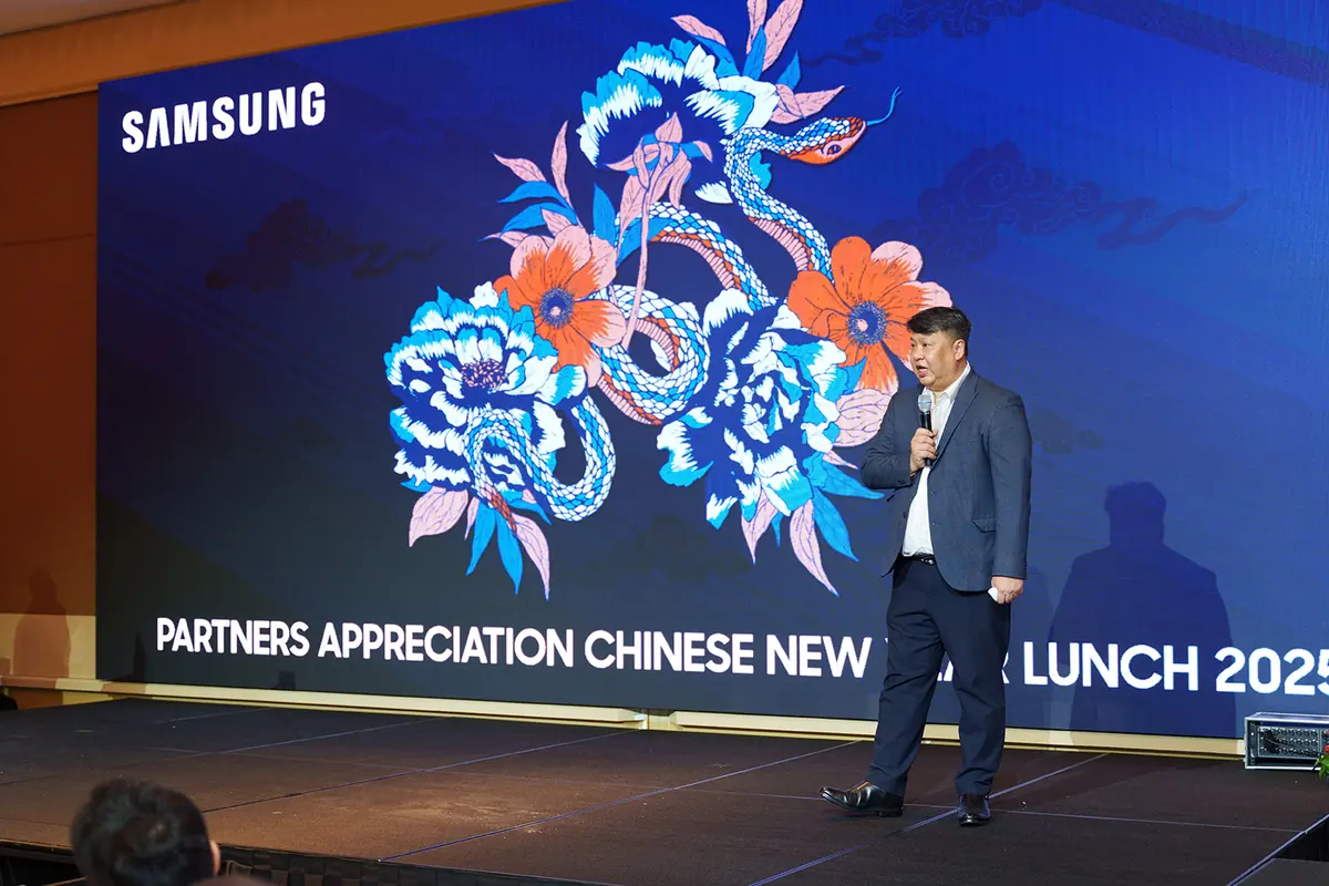 A speaker in a navy suit addresses the room from a stage, a wide screen of peonies and a coiled snake behind him