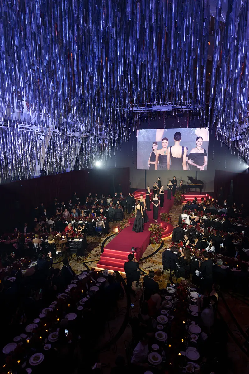A ballroom from above: hanging light strands, a red runway with models, a grand piano and long dinner tables