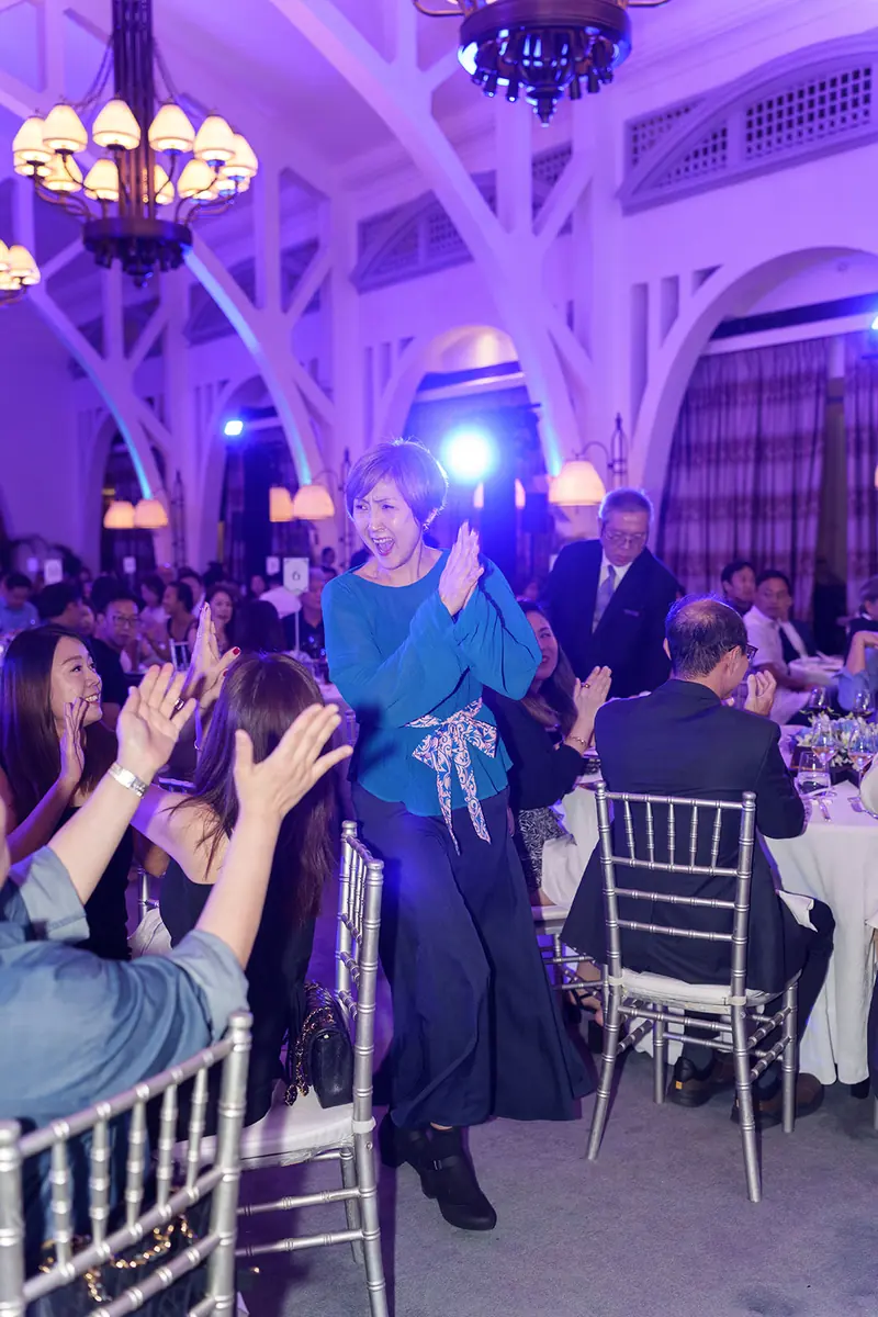 A woman in a teal top claps as she walks between round dinner tables in an arched, purple-lit hall