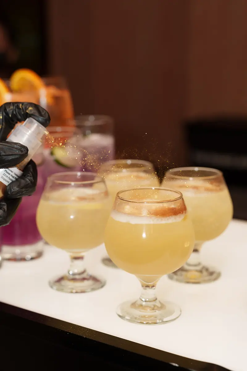 A gloved hand mists shimmer over a row of pale cocktails in stemmed glasses