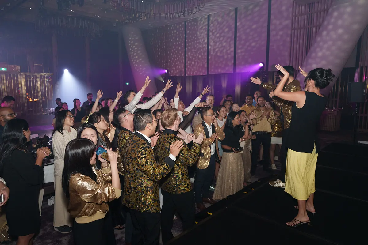 Guests in gold and black crowd the front of a stage with arms raised under purple light