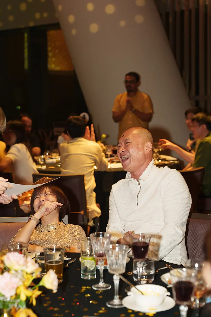A man in a cream zip top laughs at a dinner table while the woman beside him covers her face laughing
