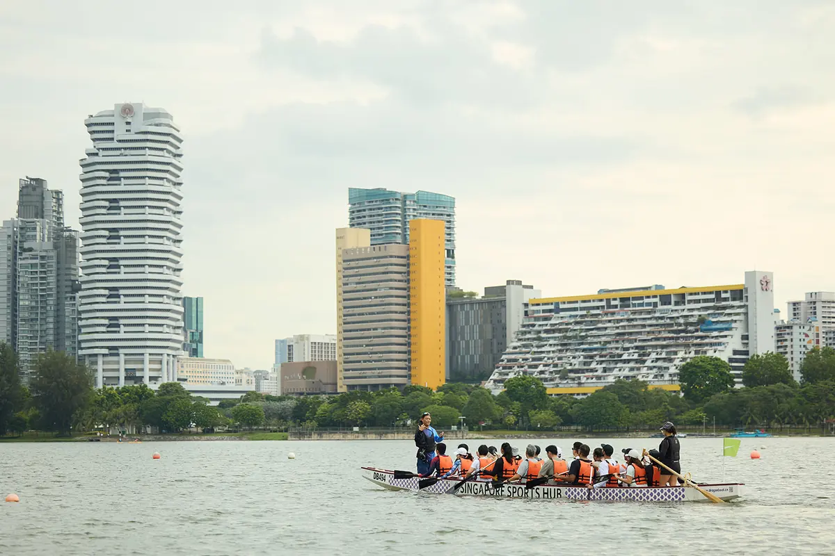 A dragon boat crew in orange life vests paddles across a wide basin below a city skyline