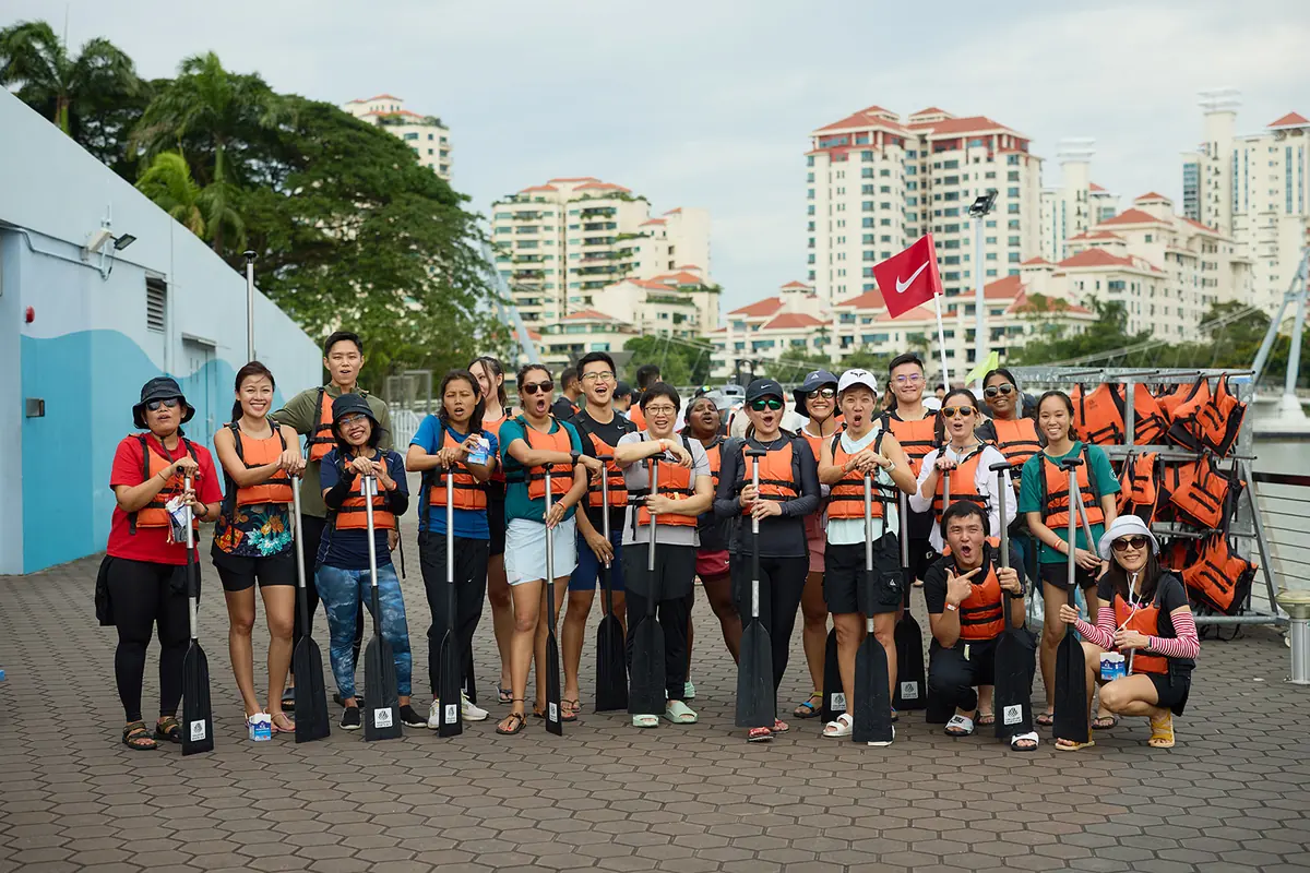 A crew in orange life vests holds paddles and poses on a jetty, apartment towers behind