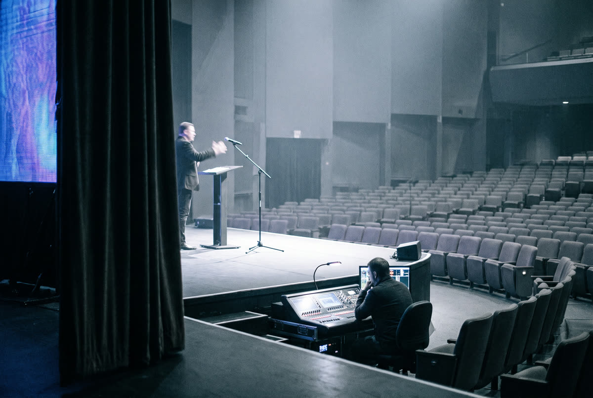 A speaker rehearsing on stage while a producer works at the control desk