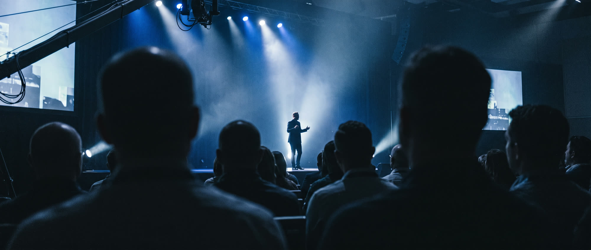 A speaker on a vast stage before a seated audience, dramatic haze and lighting